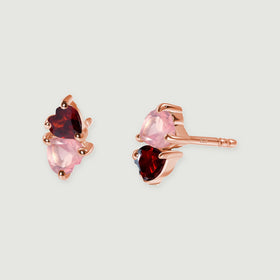 A pair of rose gold tone studs displays pale pink and deep red heart-shaped stones on a light gray surface.