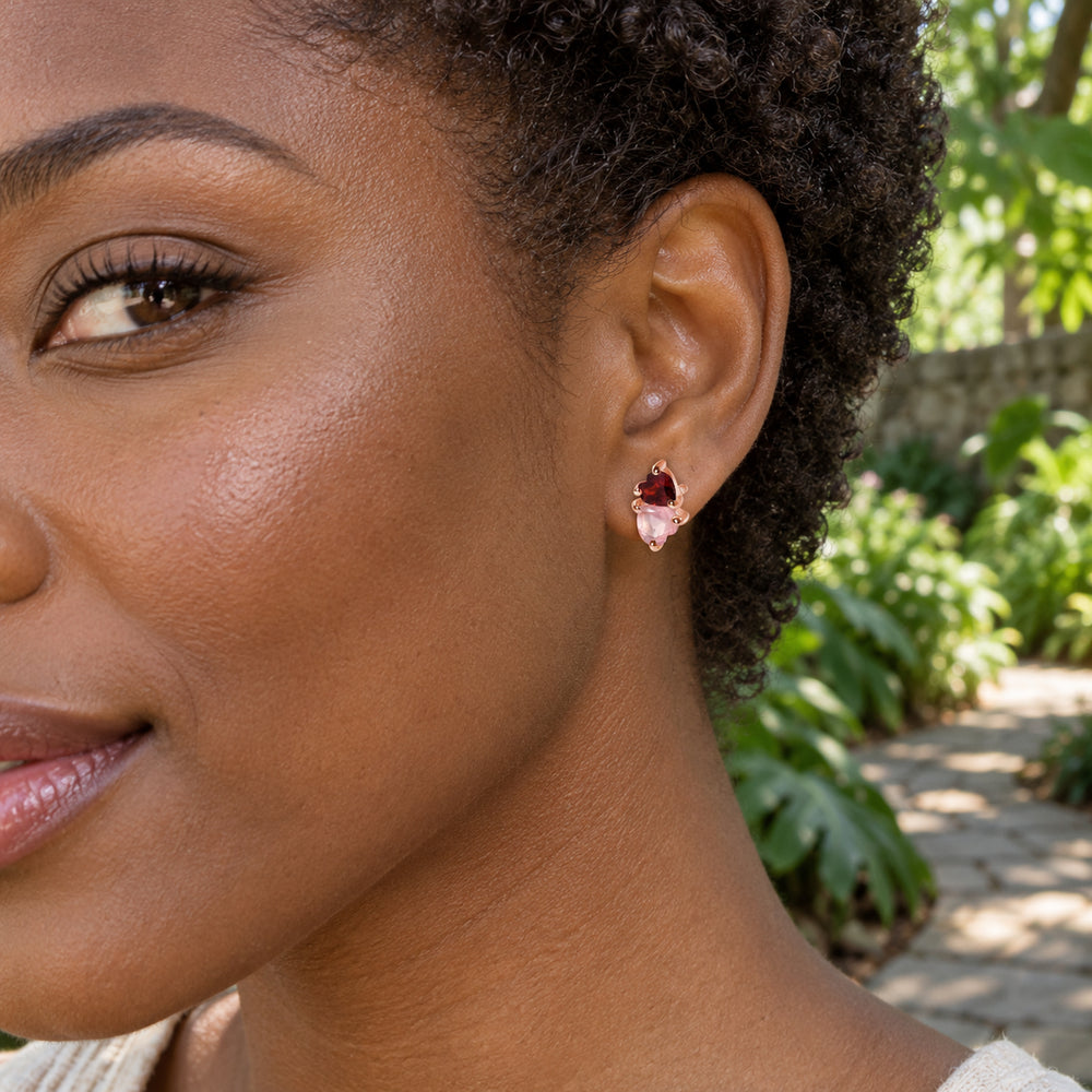 A woman with short natural curls wears a rose gold tone pink and red heart stud beside a leafy garden path.