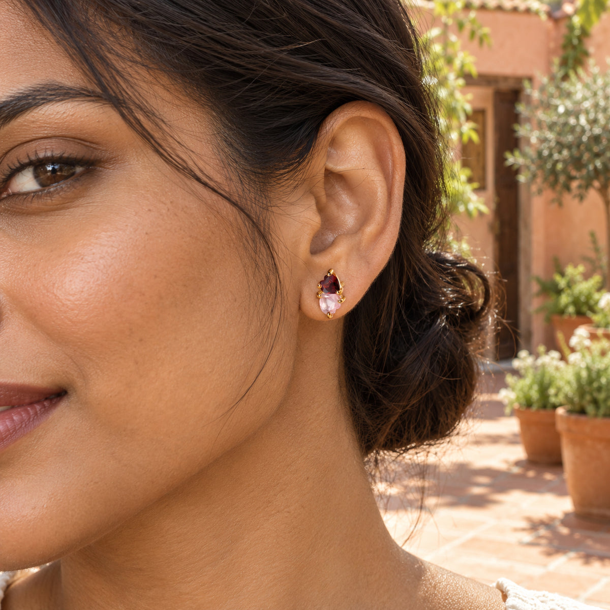 A woman with dark hair in a low bun wears a yellow gold tone two-heart stud in a sunlit terracotta courtyard.