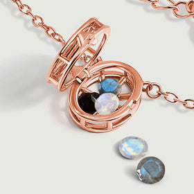 An open rose gold locket lies beside two loose faceted gemstones on a clean pale gray surface.