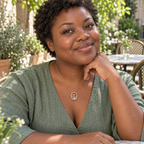 A woman with short curly hair wears the rose gold oval cage necklace with a sage linen top in a leafy courtyard cafe.