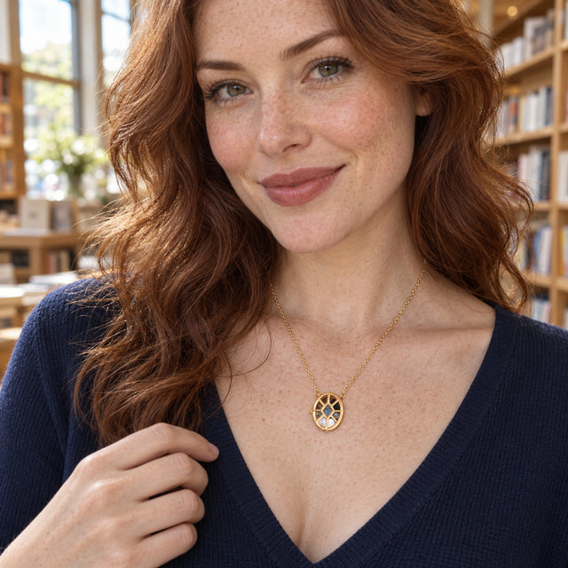A woman with wavy auburn hair wears the yellow gold gemstone locket with a navy knit top beside wooden bookshelves.