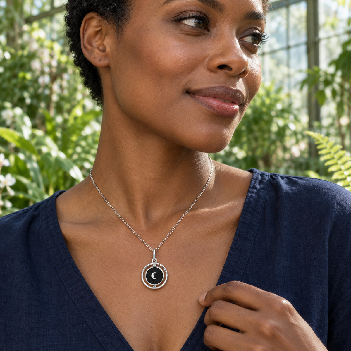 A woman in a navy blouse wears the silver black onyx crescent necklace among green plants in a glass conservatory.