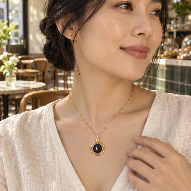 A woman in a cream linen blouse wears the yellow gold black onyx pendant in a cafe with tiled walls and wooden chairs.
