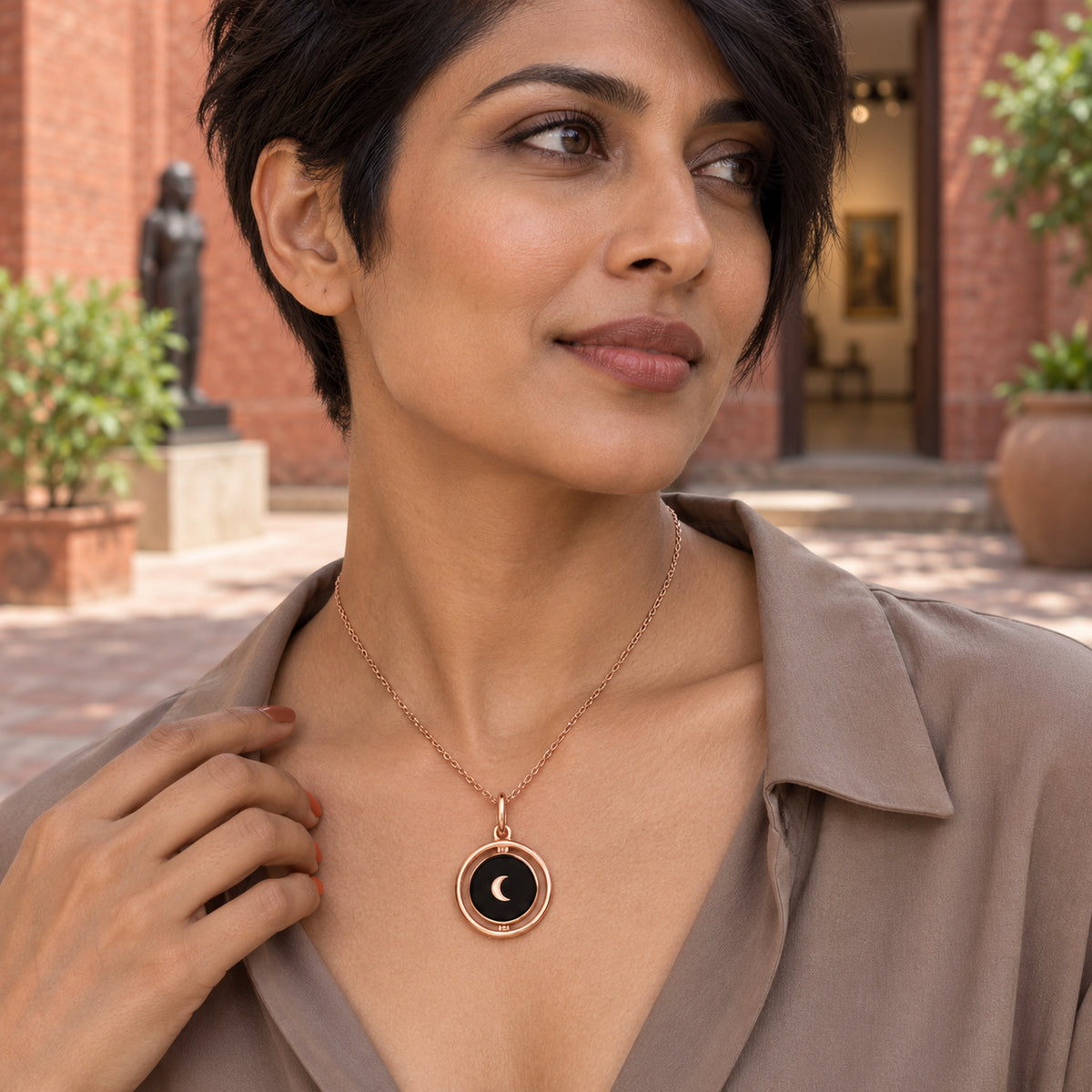 A woman with short dark hair wears the rose gold crescent necklace with a taupe blouse in a brick courtyard.