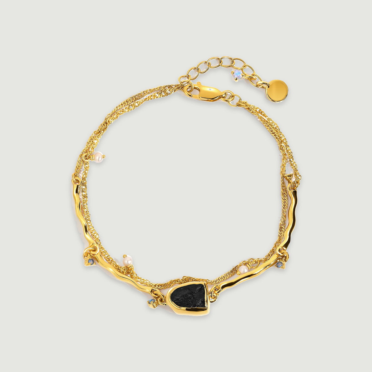 A yellow gold bracelet forms an oval with a raw black stone, small pearls, curved bars and a fine chain on a light gray surface.