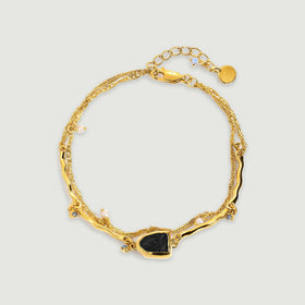 A yellow gold bracelet forms an oval with a raw black stone, small pearls, curved bars and a fine chain on a light gray surface.
