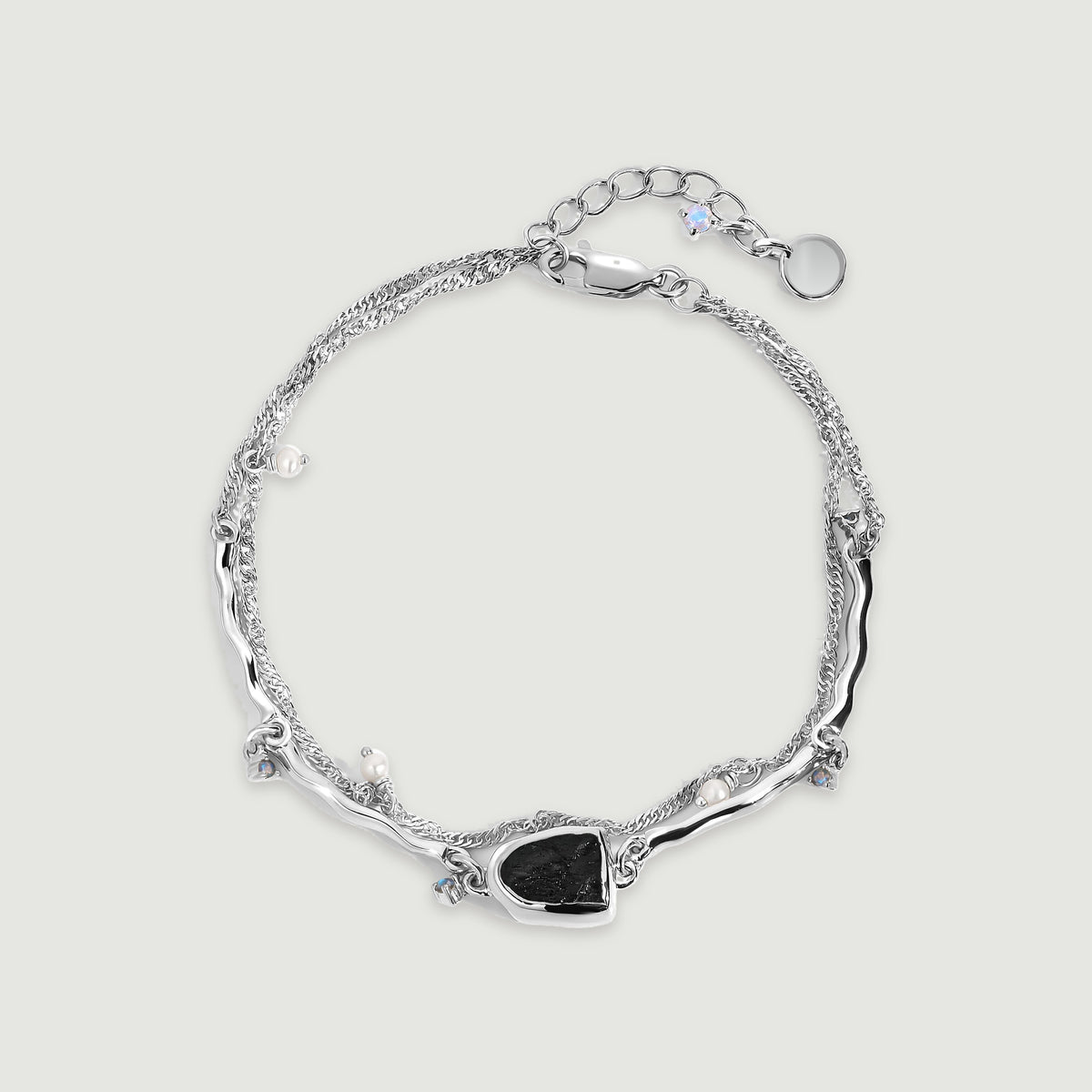 A silver bracelet is arranged in a complete loop with a dark stone centerpiece, pearl charms and an extension chain on gray.