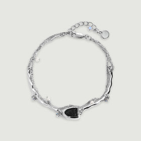 A silver bracelet is arranged in a complete loop with a dark stone centerpiece, pearl charms and an extension chain on gray.