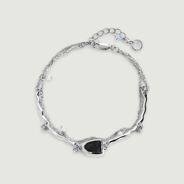 A silver bracelet is arranged in a complete loop with a dark stone centerpiece, pearl charms and an extension chain on gray.