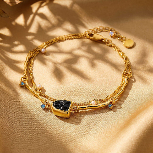A yellow gold bracelet with a black centerpiece and pearl charms lies on warm linen under soft leafy shadows.