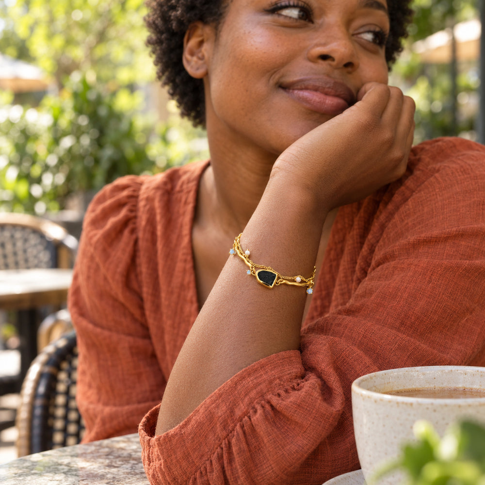 A woman in a rust linen blouse wears the yellow gold bracelet with a black stone and pearl charms at an outdoor cafe.