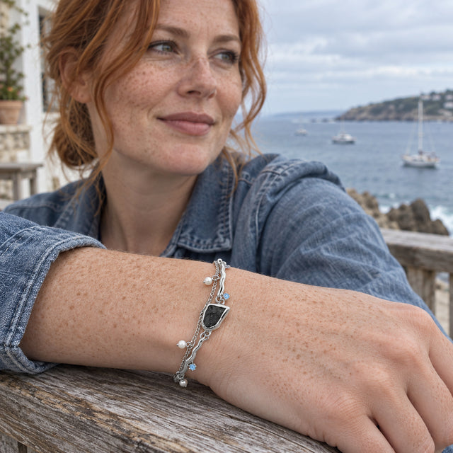 A freckled woman in a blue jacket wears the silver bracelet while resting her wrist on a wooden railing beside the sea.