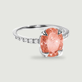 A silver ring with an oval coral peach gemstone and clear stones along the band rests on a light gray background.