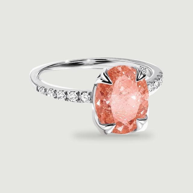 A silver ring with an oval coral peach gemstone and clear stones along the band rests on a light gray background.
