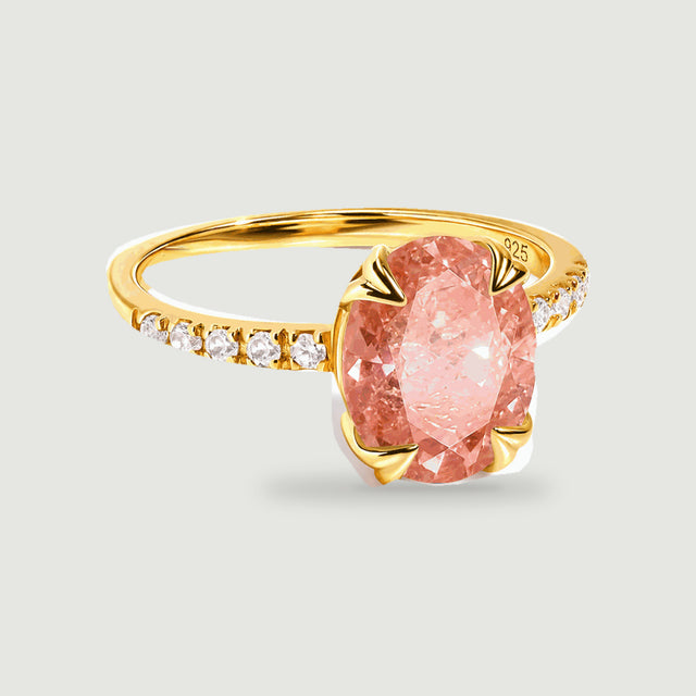 A yellow gold ring shows a faceted peach oval stone held by four heart-shaped prongs above a pale gray surface.