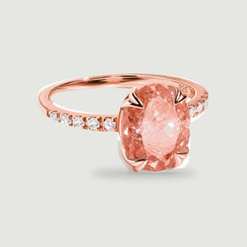 A rose gold ring with an oval peach center stone and a row of clear accents is centered on a soft gray background.