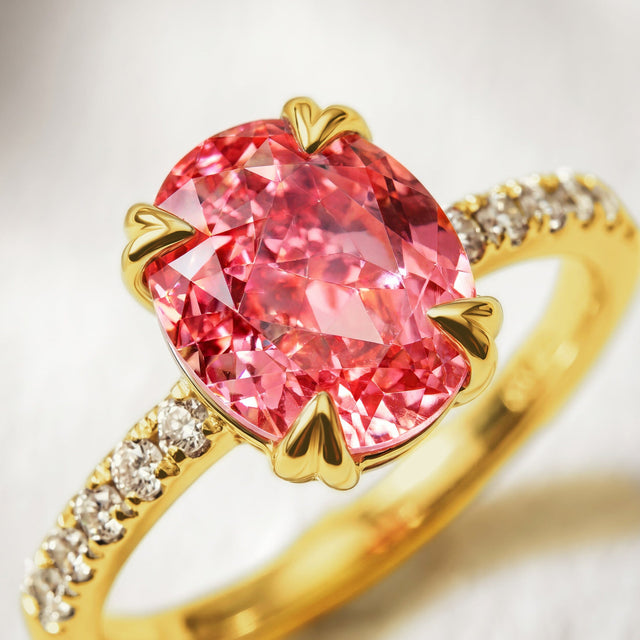 Facets of a coral pink oval stone fill the frame beside yellow gold heart-shaped prongs and round clear accents.