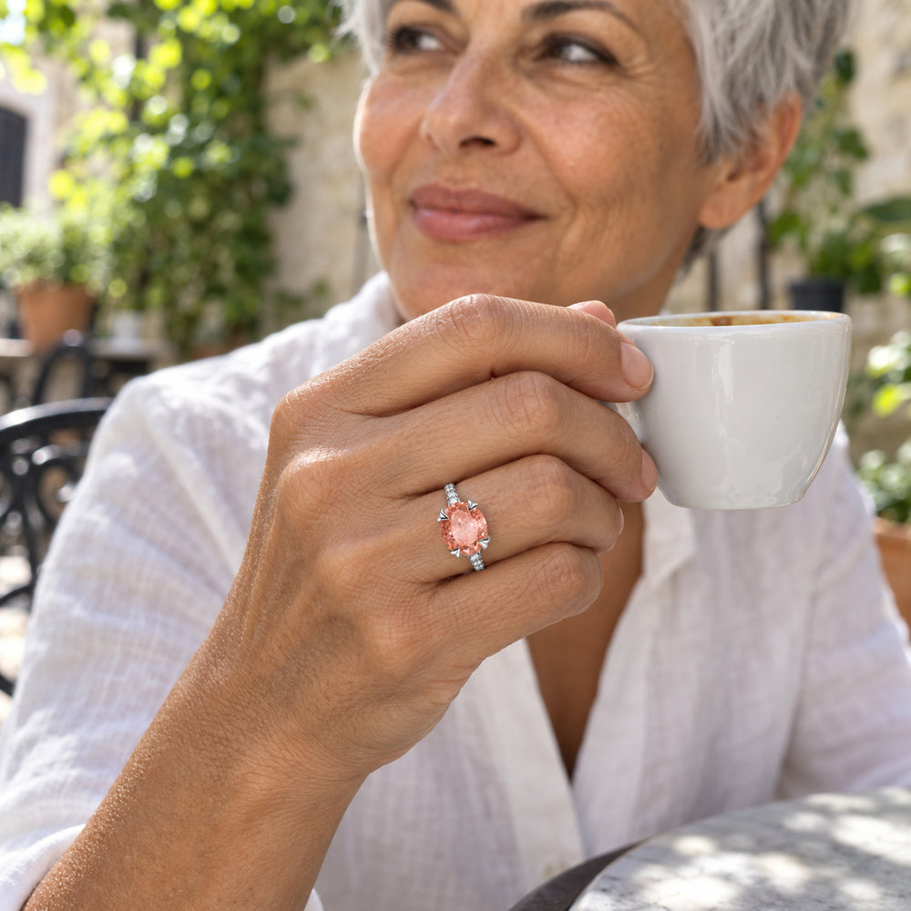 A silver ring with a peach oval gemstone and clear shoulder stones is worn on a hand holding a white cup in a leafy courtyard.