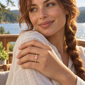 A yellow gold ring with a pink oval gemstone rests on a hand against a white linen sleeve beside a sunlit lake.