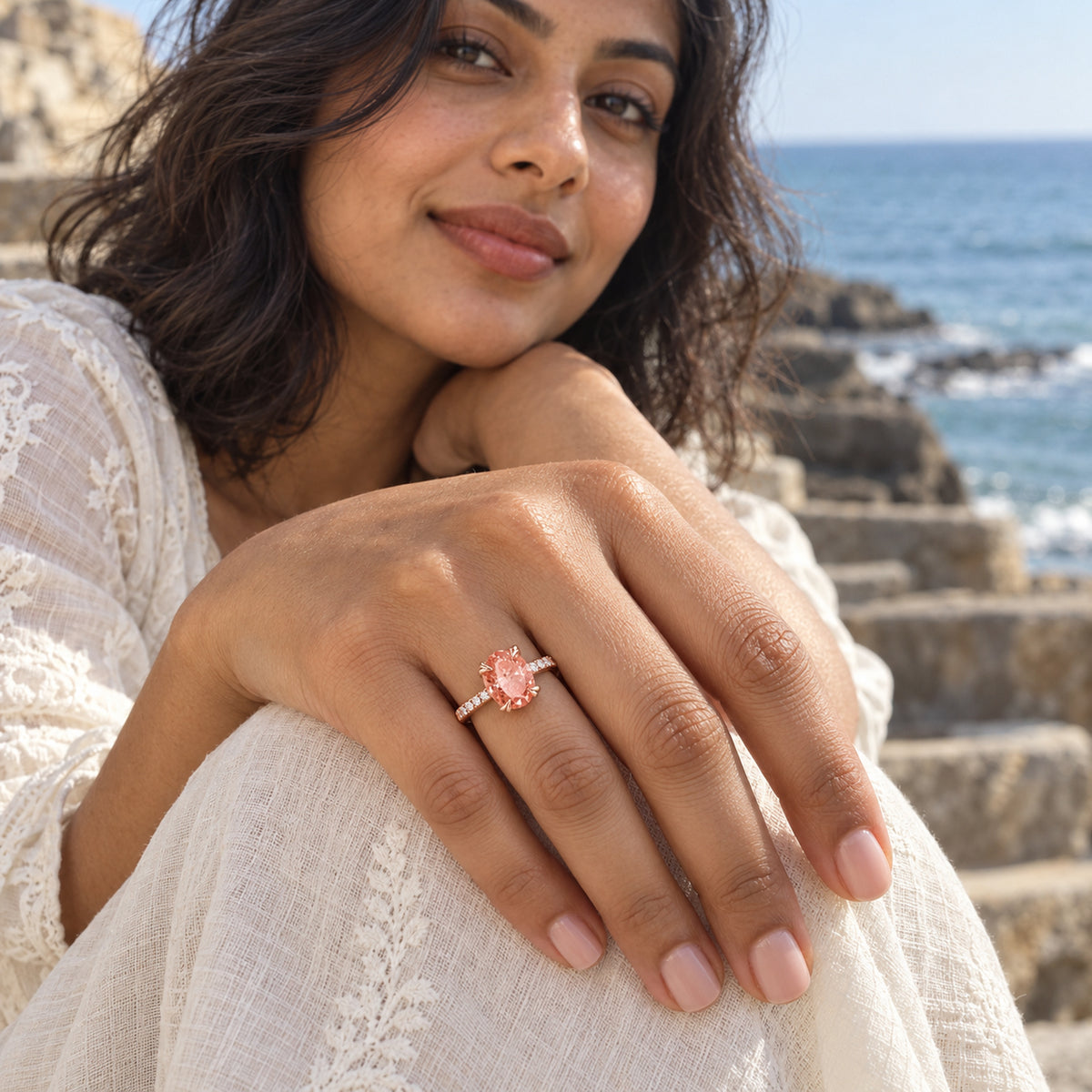 A rose gold ring with a coral peach oval stone and clear accents is shown on a hand over white fabric near the sea.