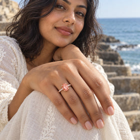 A rose gold ring with a coral peach oval stone and clear accents is shown on a hand over white fabric near the sea.