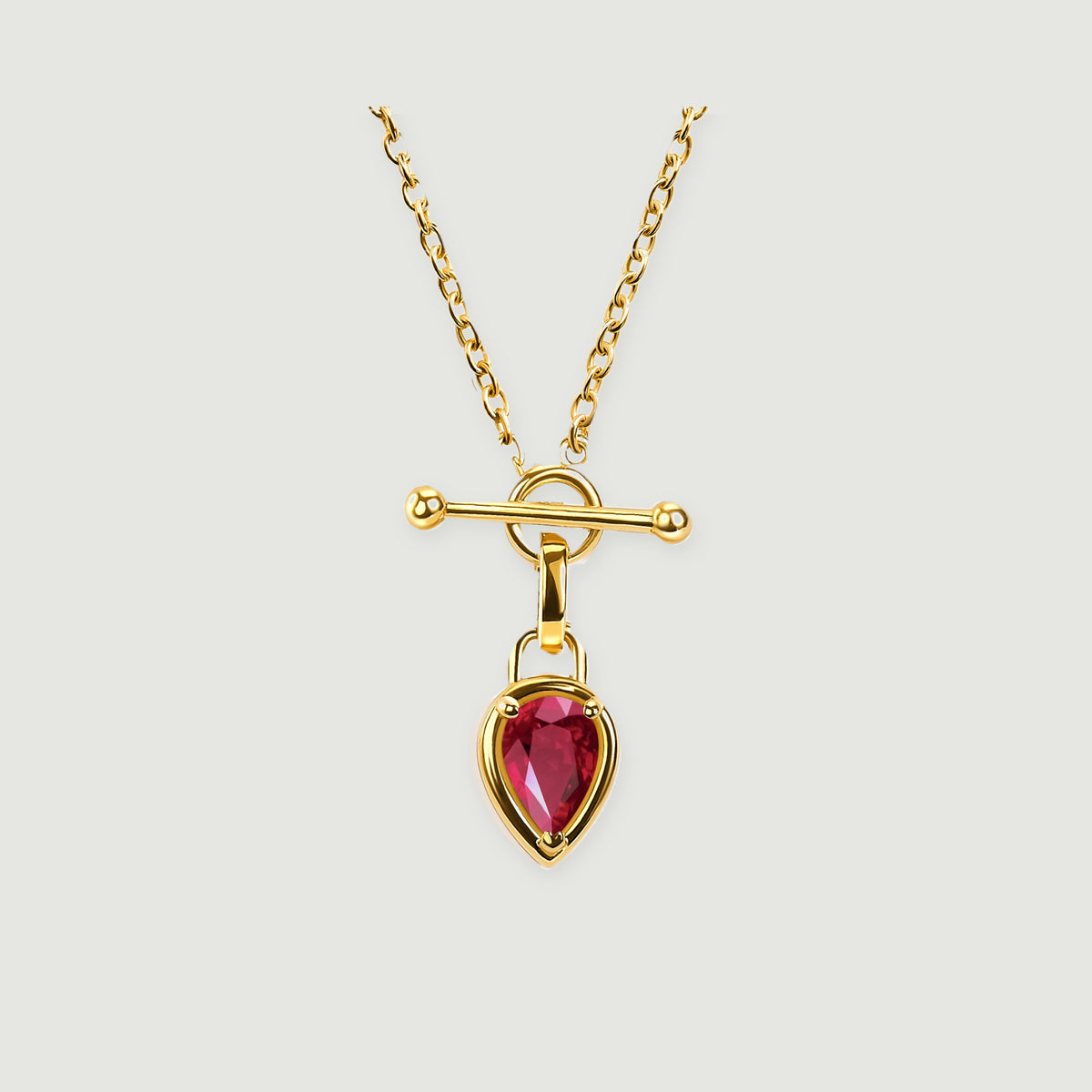A gold-tone necklace with a pear-shaped red gemstone hangs below a polished bar and circular clasp on a light gray background.