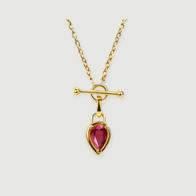 A gold-tone necklace with a pear-shaped red gemstone hangs below a polished bar and circular clasp on a light gray background.