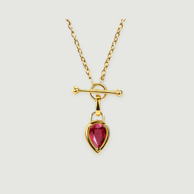 A gold-tone necklace with a pear-shaped red gemstone hangs below a polished bar and circular clasp on a light gray background.