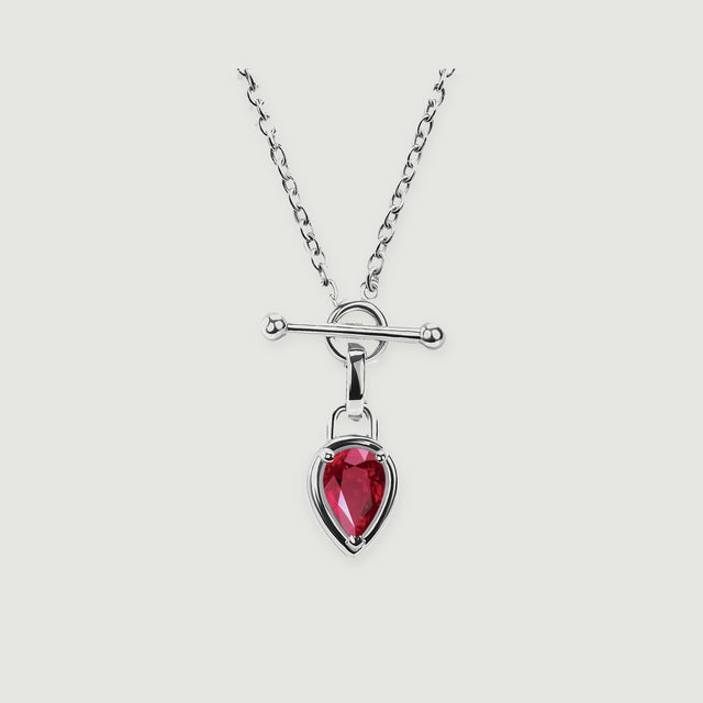 A silver-tone necklace shows a faceted red drop, an open circular clasp, and a smooth horizontal bar against pale gray.