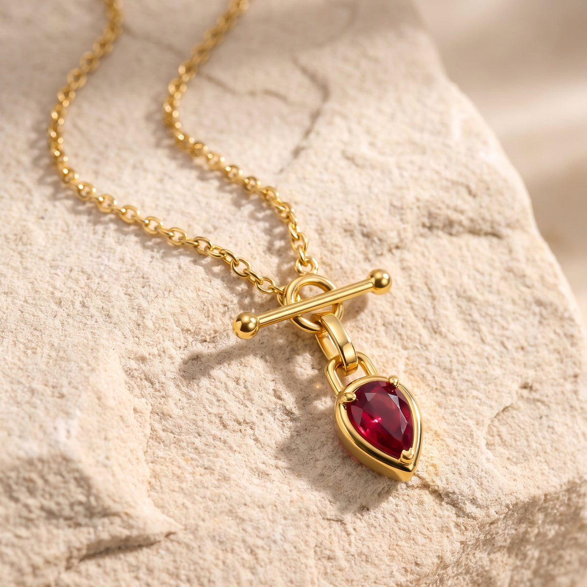 The gold-tone chain and red drop pendant rest diagonally on a textured beige stone surface.