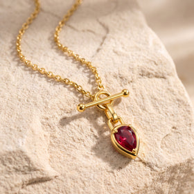 The gold-tone chain and red drop pendant rest diagonally on a textured beige stone surface.