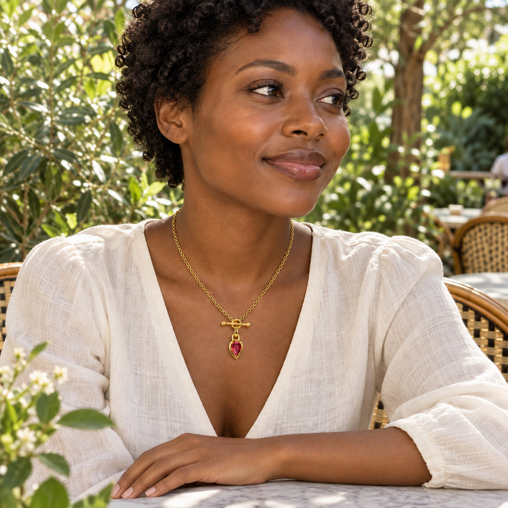 A woman with short curls wears the gold-tone ruby necklace with an ivory blouse at a leafy outdoor cafe.