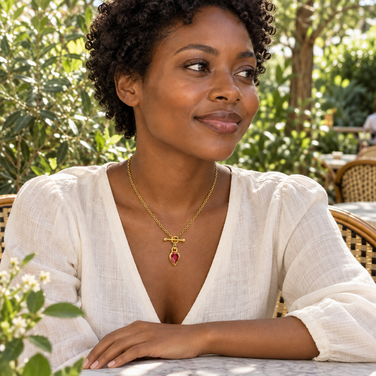 A woman with short curls wears the gold-tone ruby necklace with an ivory blouse at a leafy outdoor cafe.