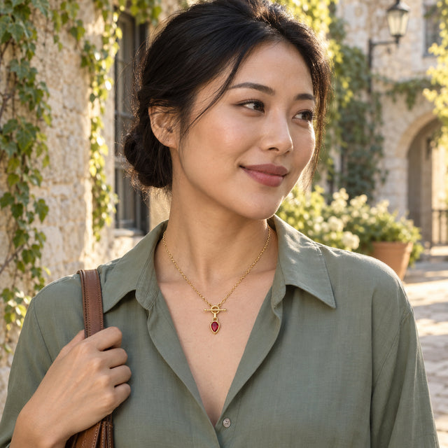The gold-tone ruby pendant is visible above a sage green blouse as a woman holds a bag strap in a sunny stone courtyard.