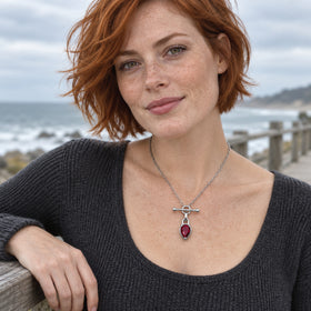 A red-haired woman wears the silver-tone red gemstone necklace with a charcoal knit top beside a coastal railing.