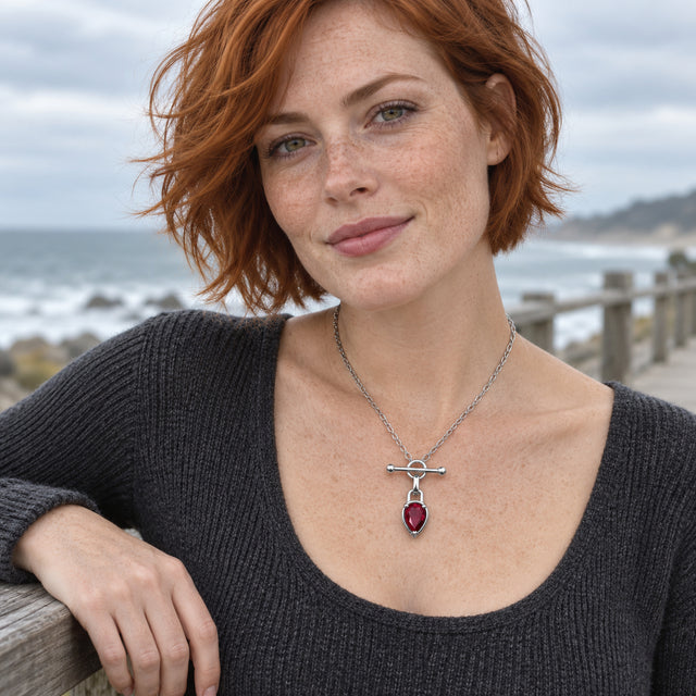A red-haired woman wears the silver-tone red gemstone necklace with a charcoal knit top beside a coastal railing.