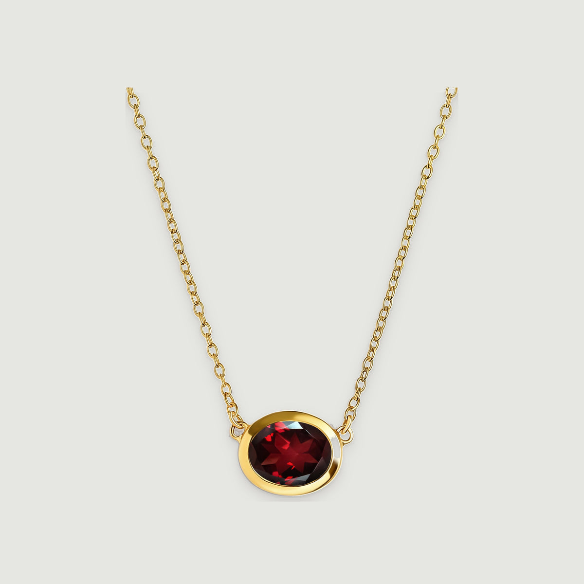 A yellow gold plated necklace with an oval faceted garnet is centered above a soft shadow on light grey.