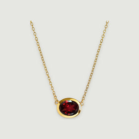 A yellow gold plated necklace with an oval faceted garnet is centered above a soft shadow on light grey.