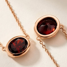Two close views show the dark red facets, polished rose gold plated bezel, and small chain links on a pale textured surface.