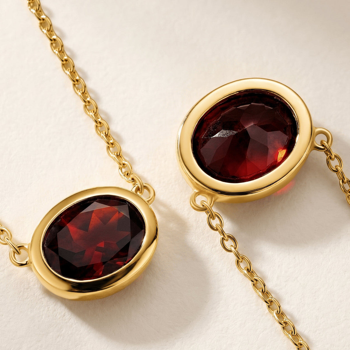 The front and angled faces of an oval garnet are shown with yellow gold plated rims and link chains on a cream surface.