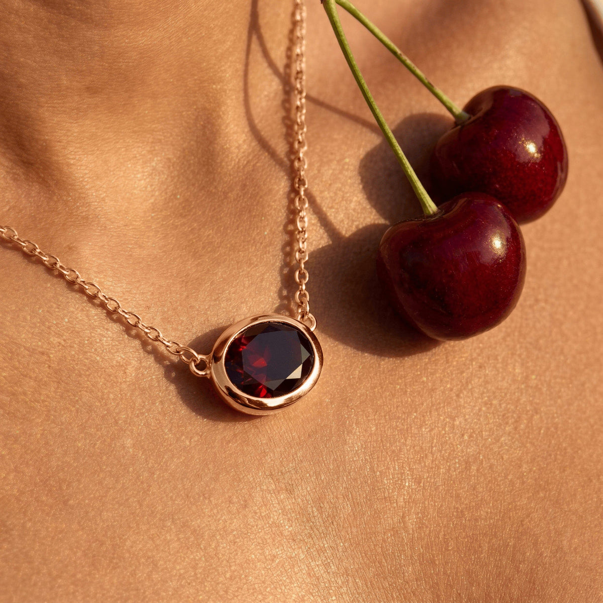 A rose gold plated garnet necklace rests beside two red cherries on a warm textured surface.