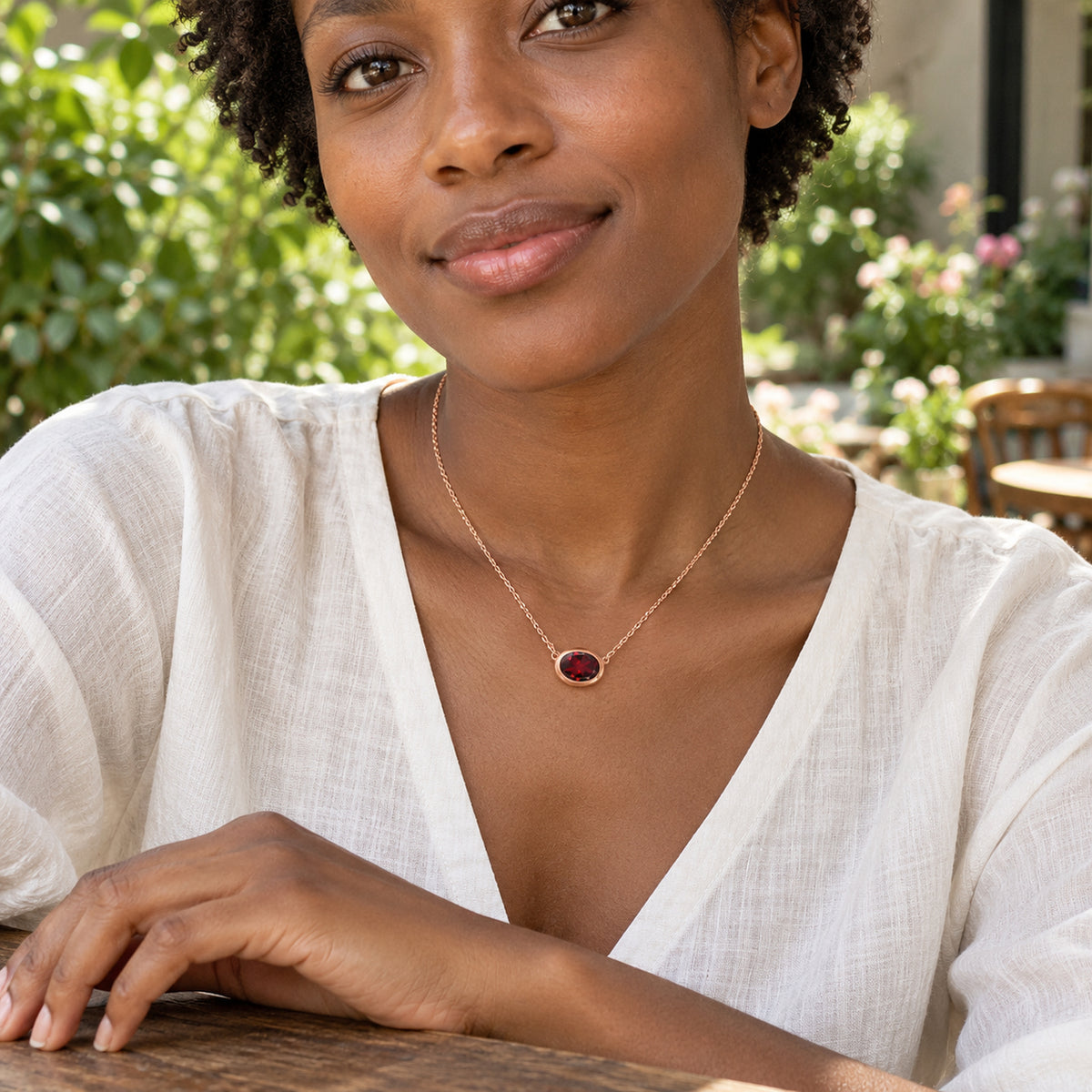 A woman in an ivory linen blouse wears the rose gold plated garnet necklace at an outdoor cafe with leafy greenery behind her.