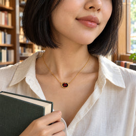 A woman with a black bob wears the yellow gold plated garnet necklace while holding a green book beside a sunlit bookstore window.