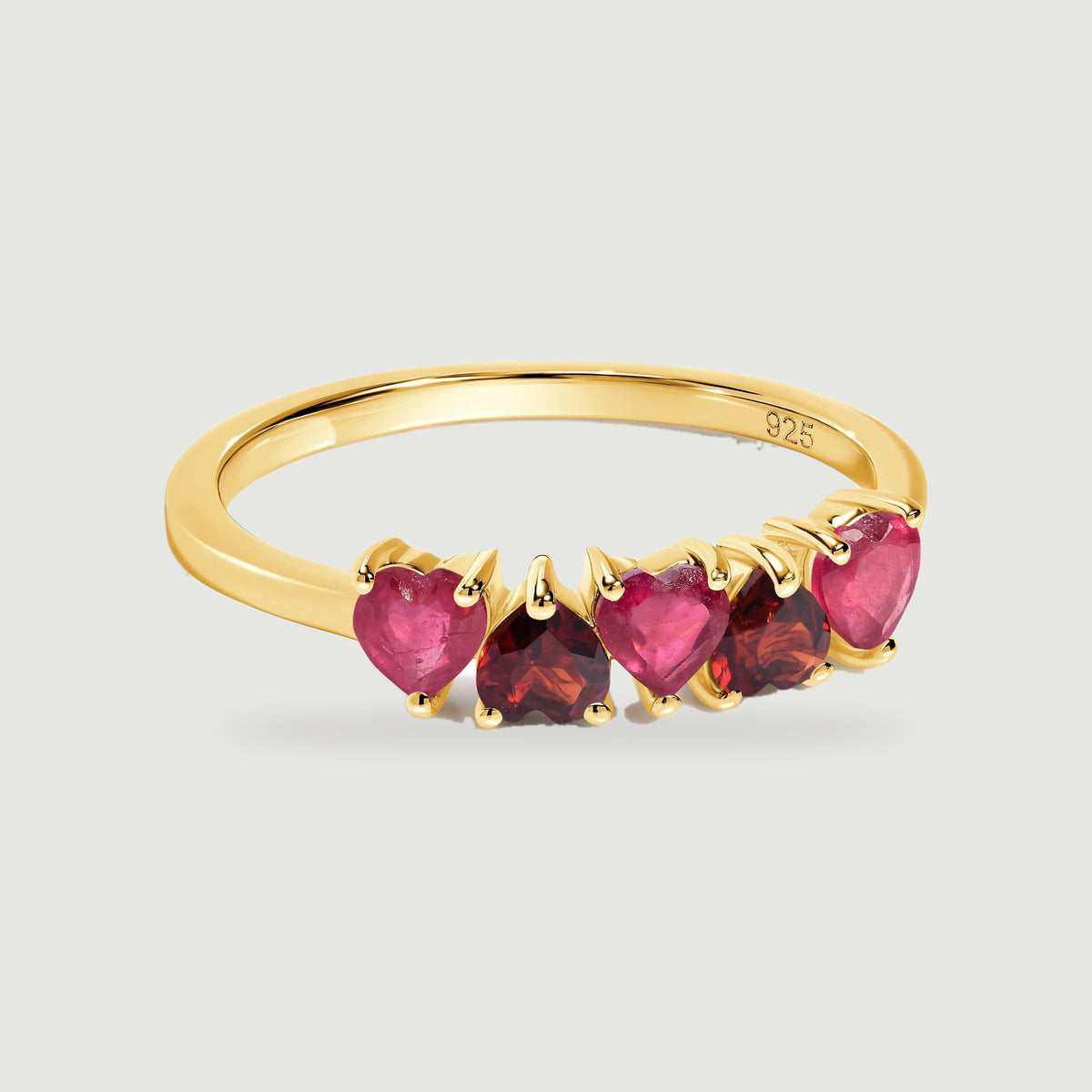 A yellow gold ring displays five alternating pink and dark red heart-shaped stones on a light gray background.