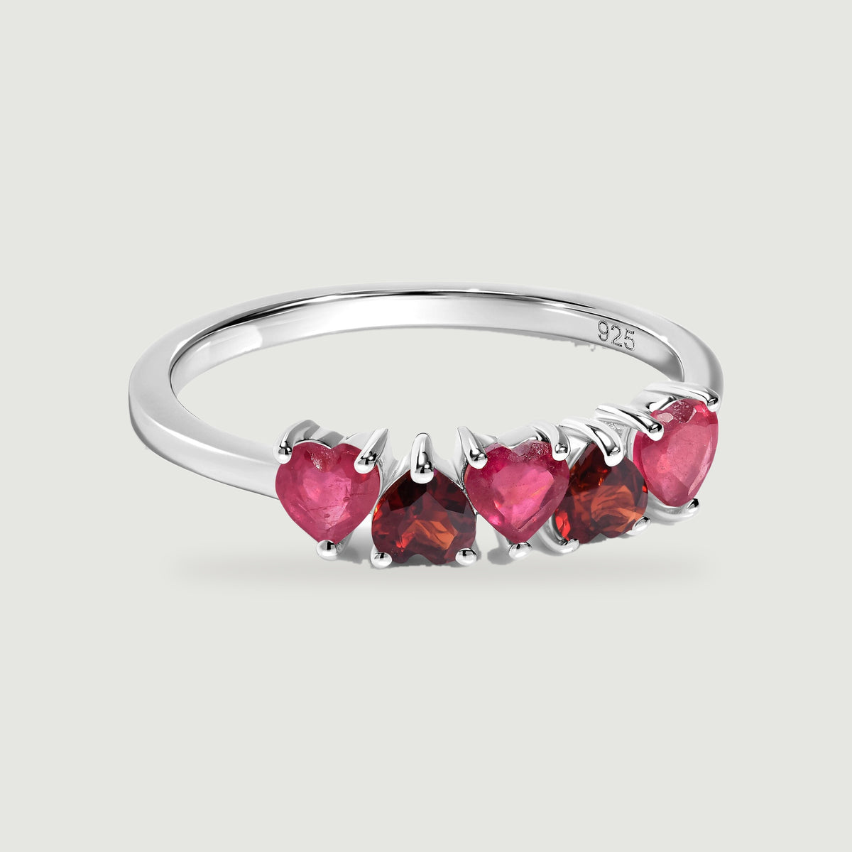 A silver ring with polished prongs holds a row of pink and deep red heart-shaped gemstones.