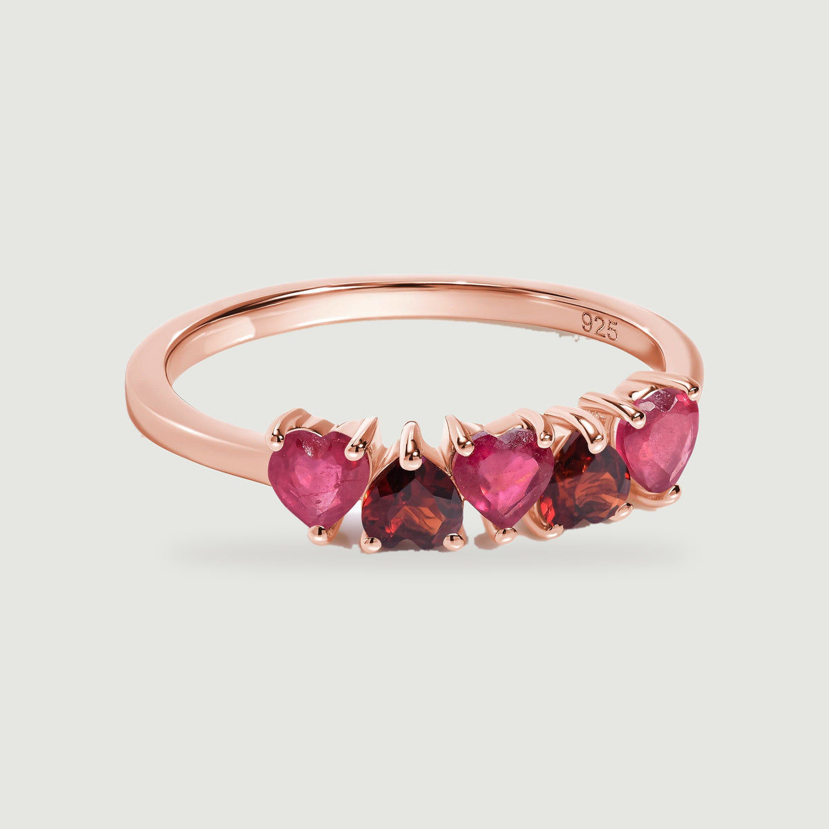 A rose gold ring with five heart-shaped stones is centered above a soft shadow on a light gray surface.