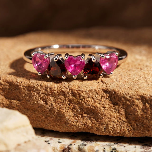 Pink ruby and dark red garnet hearts line the silver ring resting on a warm stone slab.