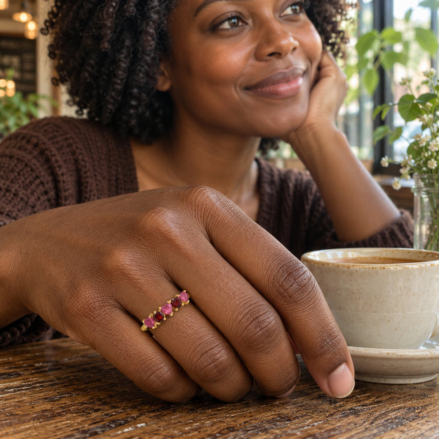 A yellow gold heart-stone ring is worn on a hand resting beside a ceramic coffee cup in a cafe.