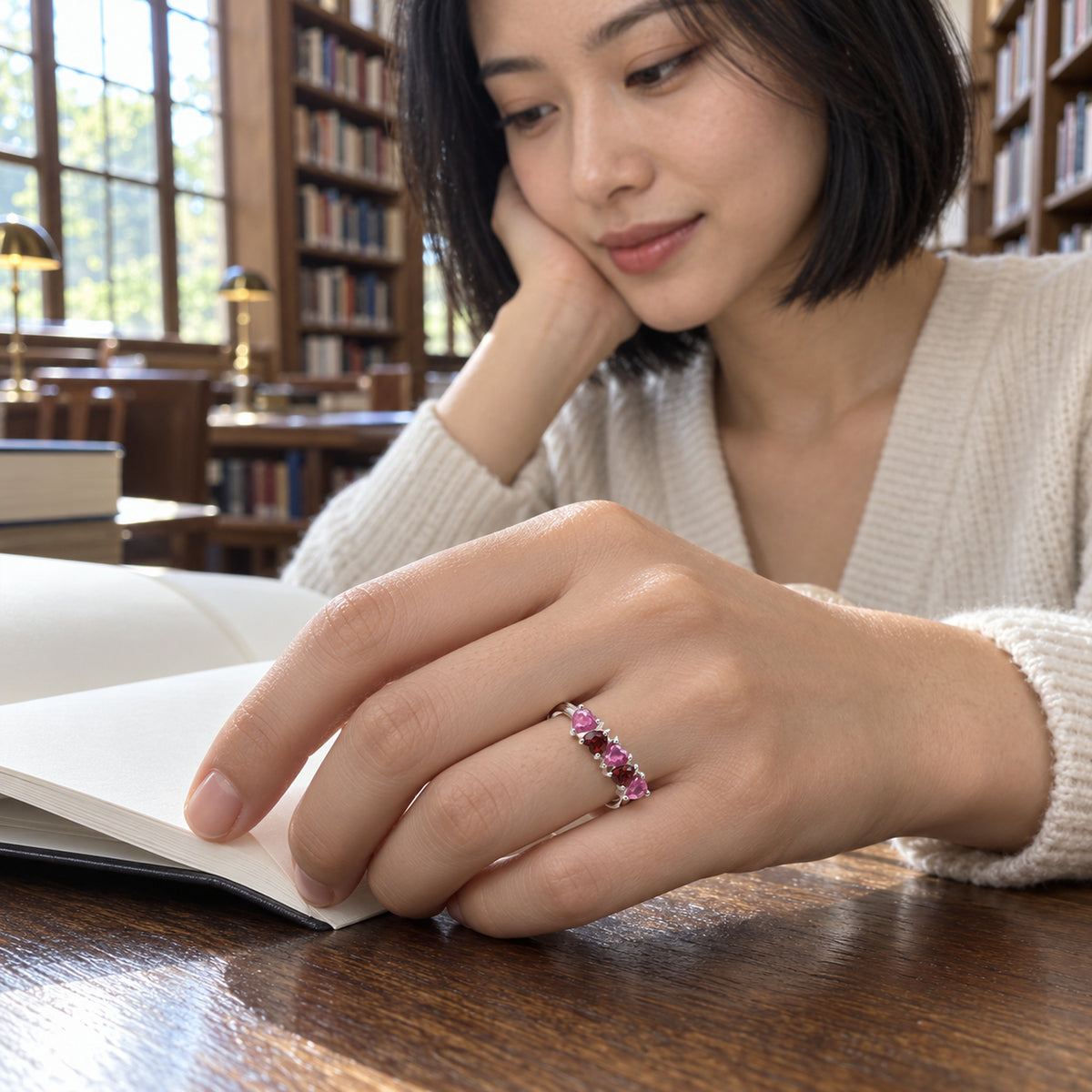 A silver ring with pink and wine-red heart stones is shown on a hand resting on an open book in a library.